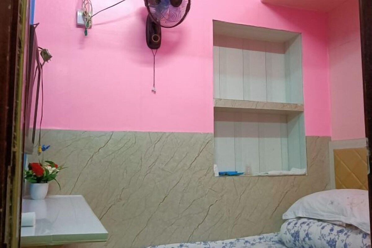 Quarto com parede rosa, parte inferior com padrão marmorizado, mesa de cabeceira com uma planta, cama com travesseiro floral e uma pequena janela com prateleira acima.