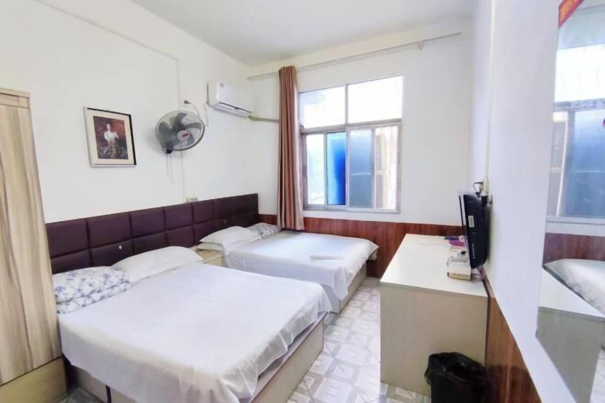 Ein Hotelzimmer mit zwei Betten, einem Schreibtisch, einem Stuhl, einem Fernseher, einem Fenster mit Vorhängen und einem Wandventilator.