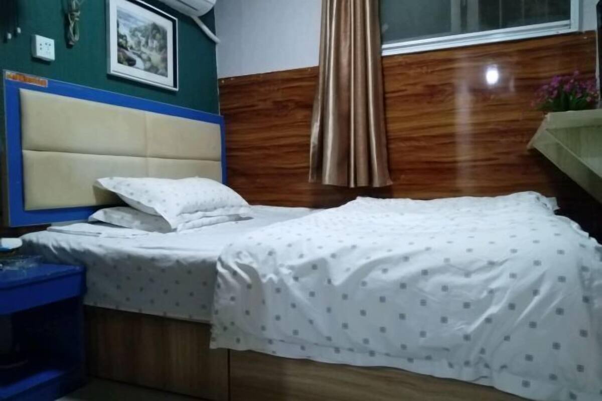 Ein Schlafzimmer mit einem Bett, einem blauen Nachttisch, einem Holz-Kopfteil, einem Fenster mit Vorhängen und einer Wand-Klimaanlage.