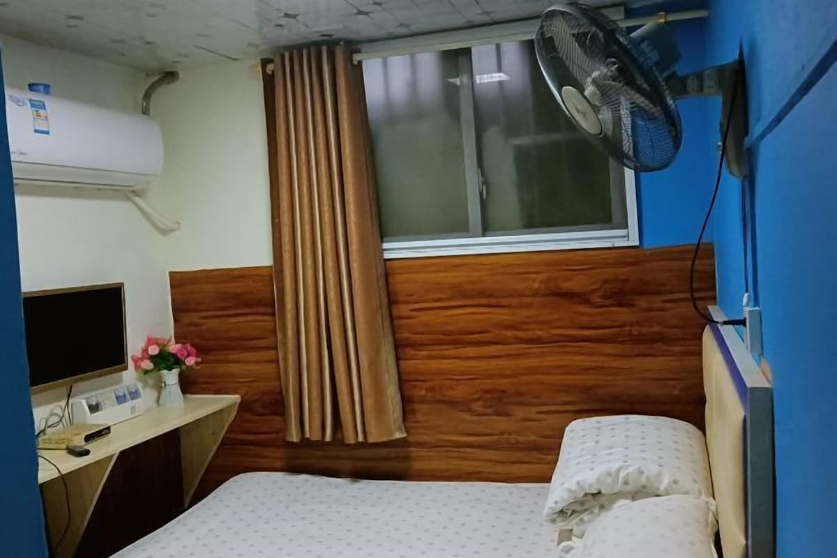 Quarto de hotel com cama, escrivaninha com computador, ventilador fixado na parede, janela com cortinas e aparelho de ar condicionado.
