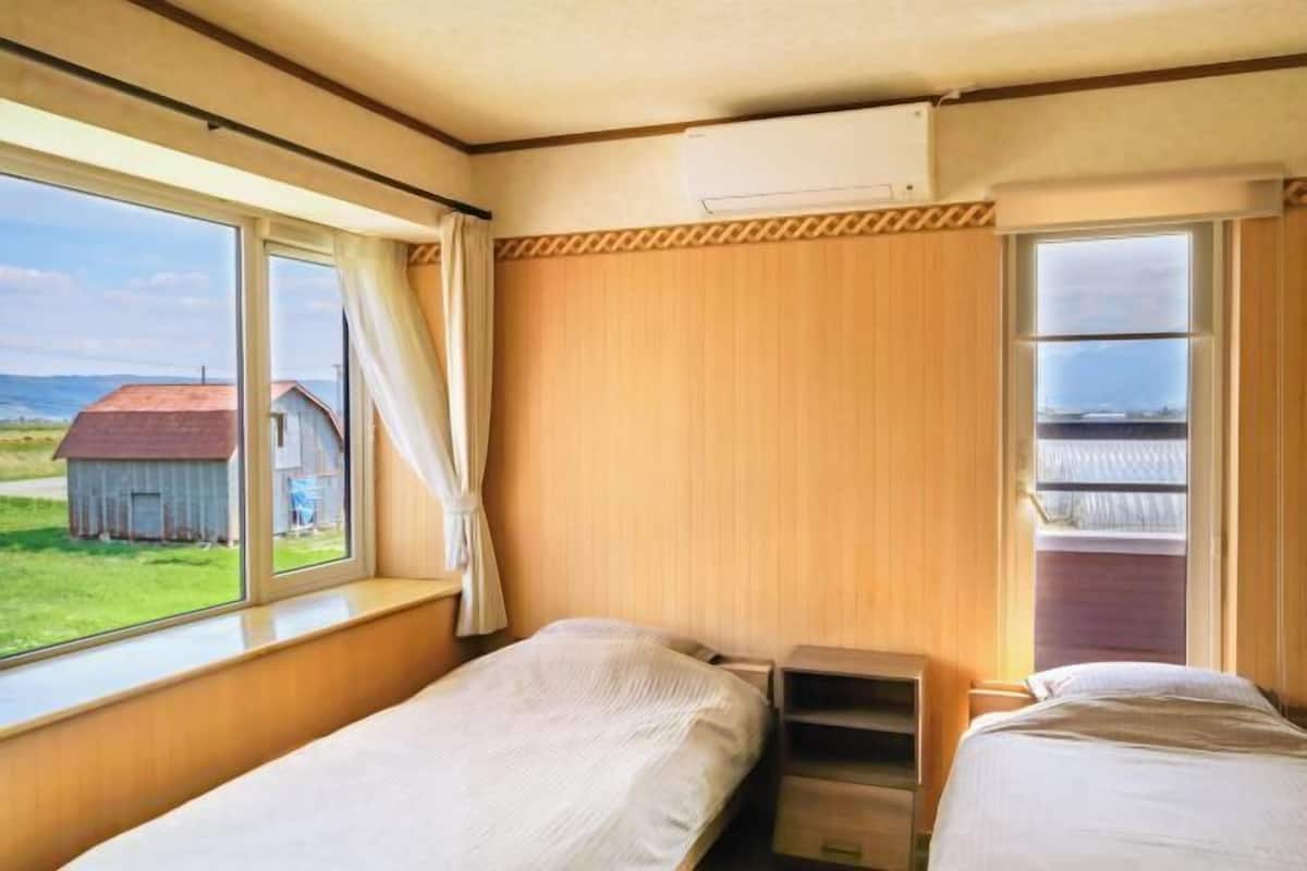 A room with two beds, a window with a view of a rural landscape, and an air conditioning unit on the wall.