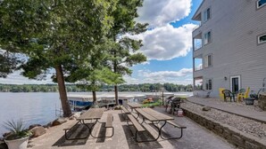 Eine Terrasse am Seeufer mit Picknicktischen, einer Grillgelegenheit und einem Gebäude mit Balkon mit Blick auf das Wasser.