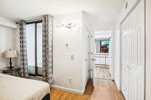 A hotel room with a bed, a nightstand with a lamp, a patterned curtain, a sliding door leading to a kitchen, and a hallway with wooden flooring. - Dells Lake Lookout At Lighthouse Cove Condominiums (Wisconsin Dells)