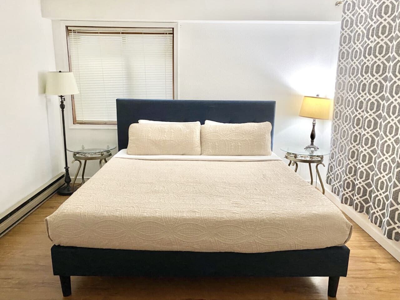 Una cama bien tendida con un cabecero oscuro, una mesita de noche con lámpara y un papel tapiz estampado.