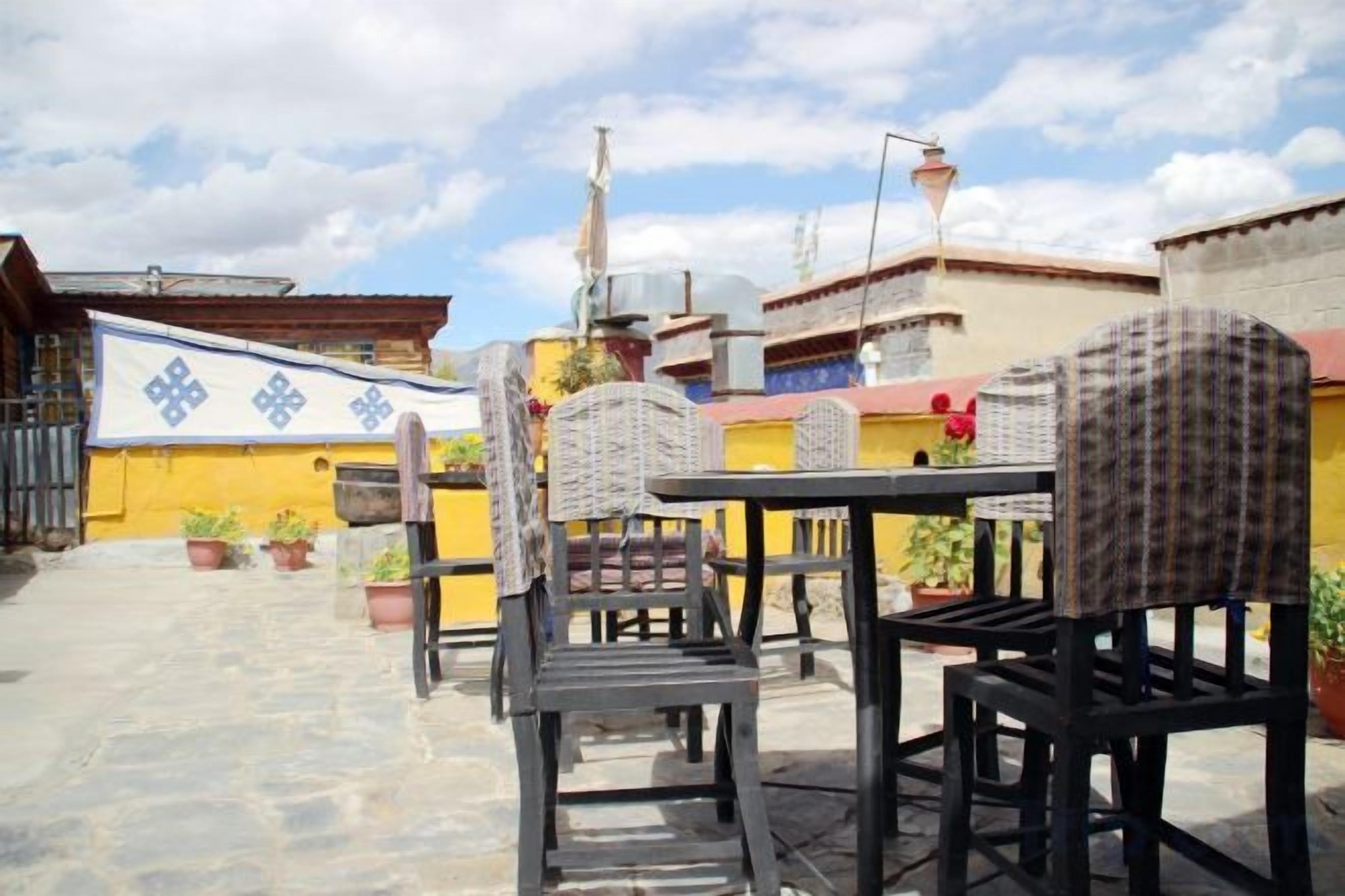 An outdoor patio area with tables and chairs, potted plants, and a yellow building in the background.