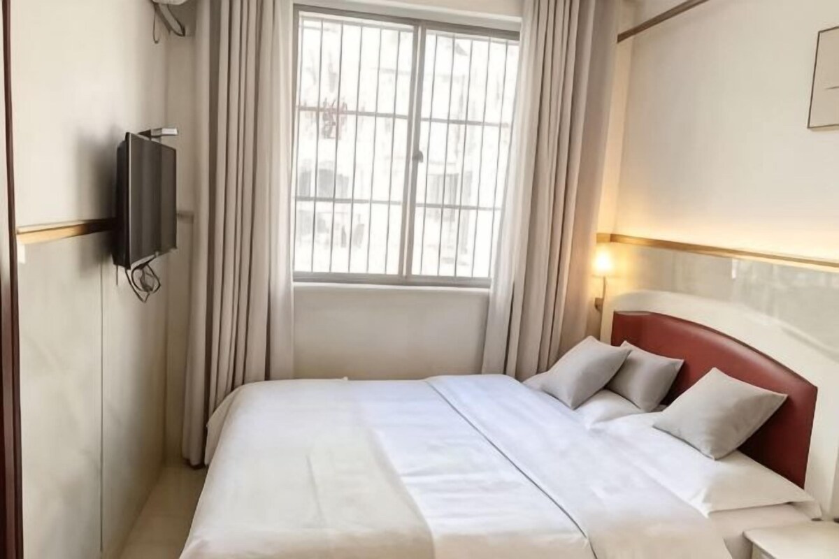 Une chambre d’hôtel avec un lit, un téléviseur fixé au mur, une fenêtre avec des rideaux et une lampe de chevet.