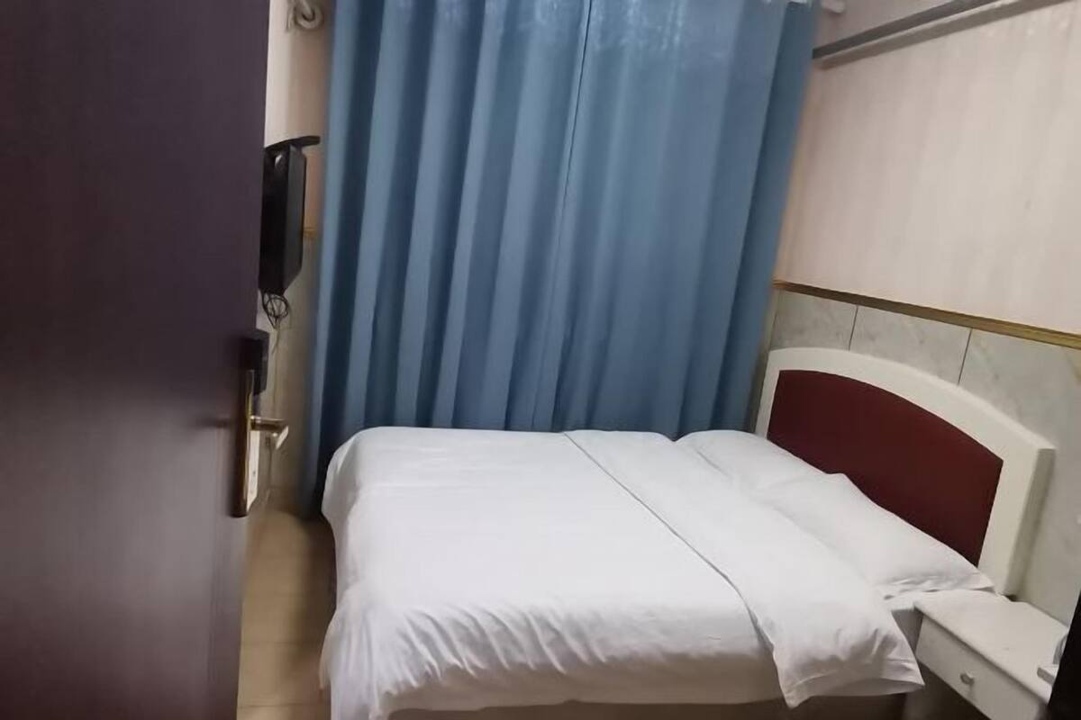 Een kleine kamer met een enkel bed, een witte spreidek, een rood hoofdeinde en een blauw gordijn.