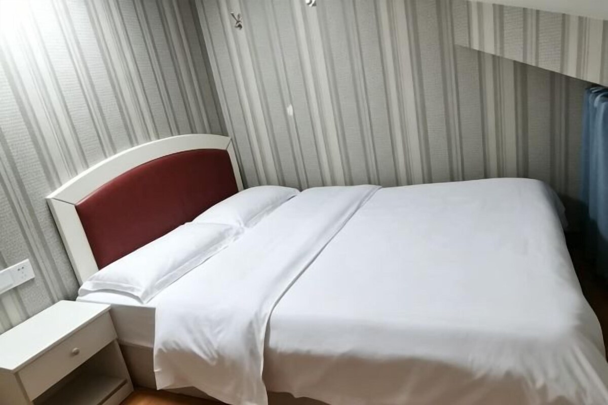 Un lit simple avec un matelas blanc et une tête de lit rouge, une table de chevet avec un tiroir et un mur à rayures.