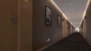 Um corredor de hotel com obras de arte emolduradas nas paredes, piso coberto por carpete, e um longo caminho até uma porta no final.