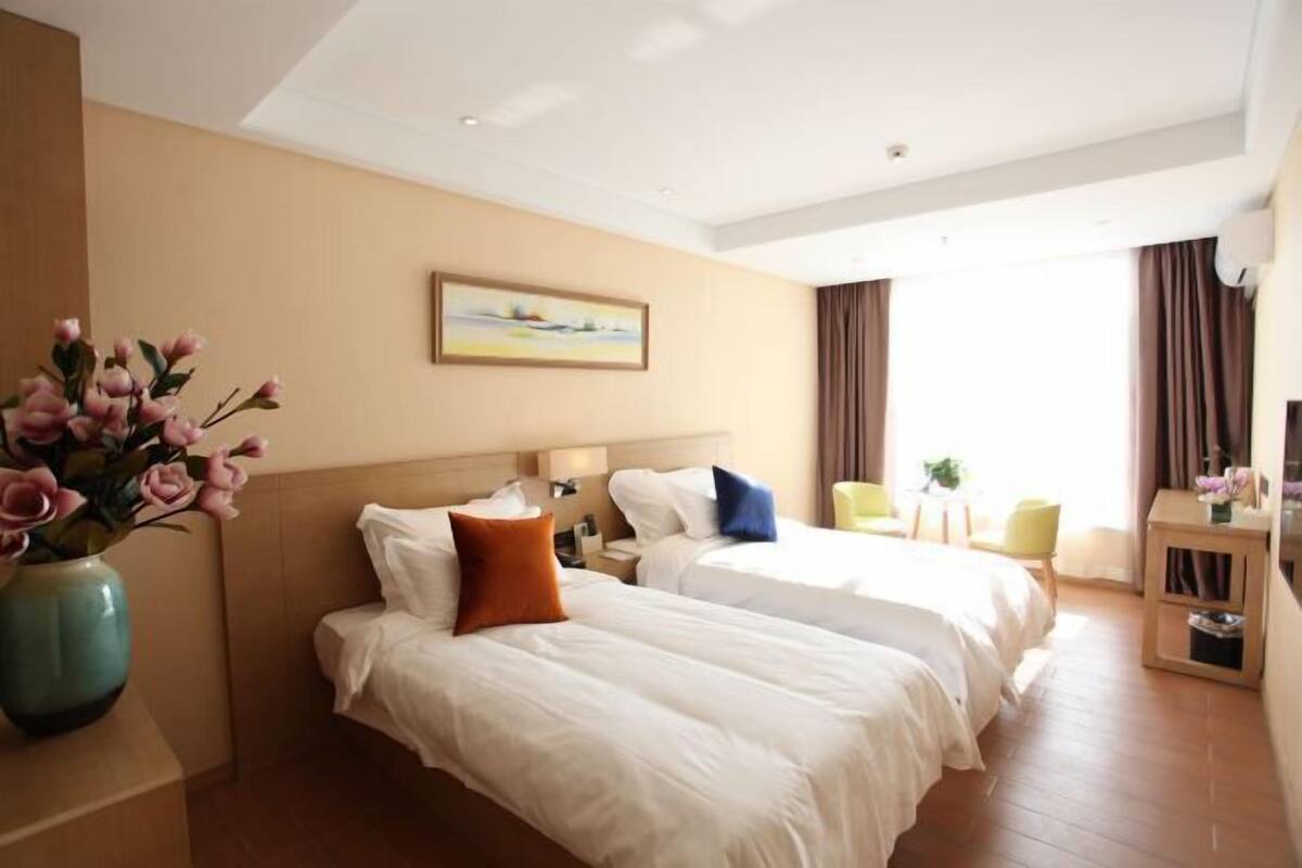 A hotel room with two beds, a vase of flowers, a painting on the wall, and a view of the outdoors.