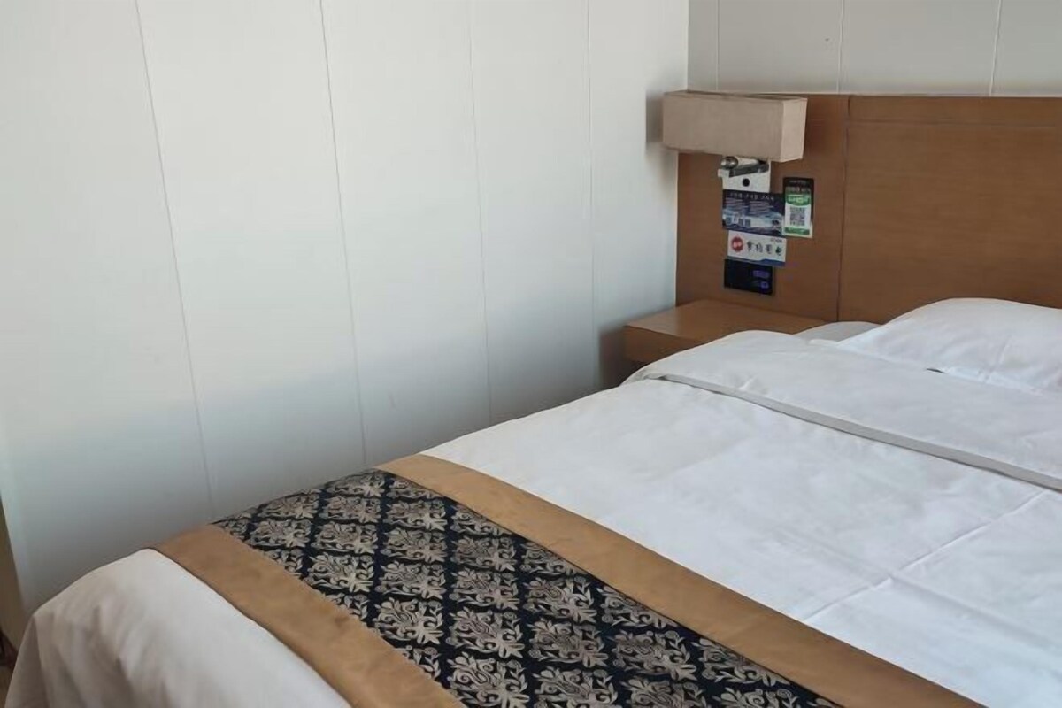 Quarto de hotel com cama, mesa de cabeceira, luminária na parede e um mural com avisos.