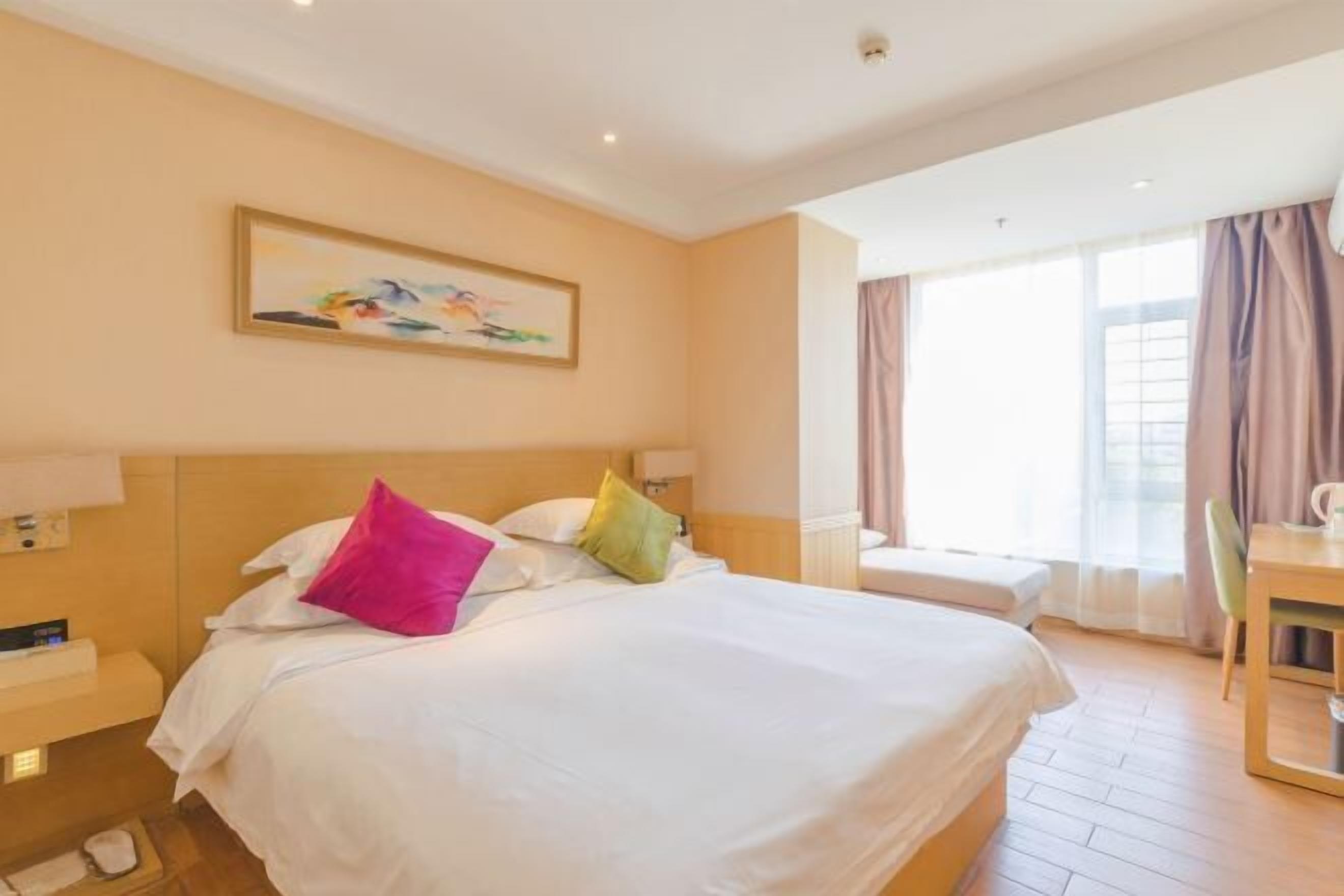 A hotel room with a large bed, a bedside table, a chair, a window with curtains, and a painting on the wall.