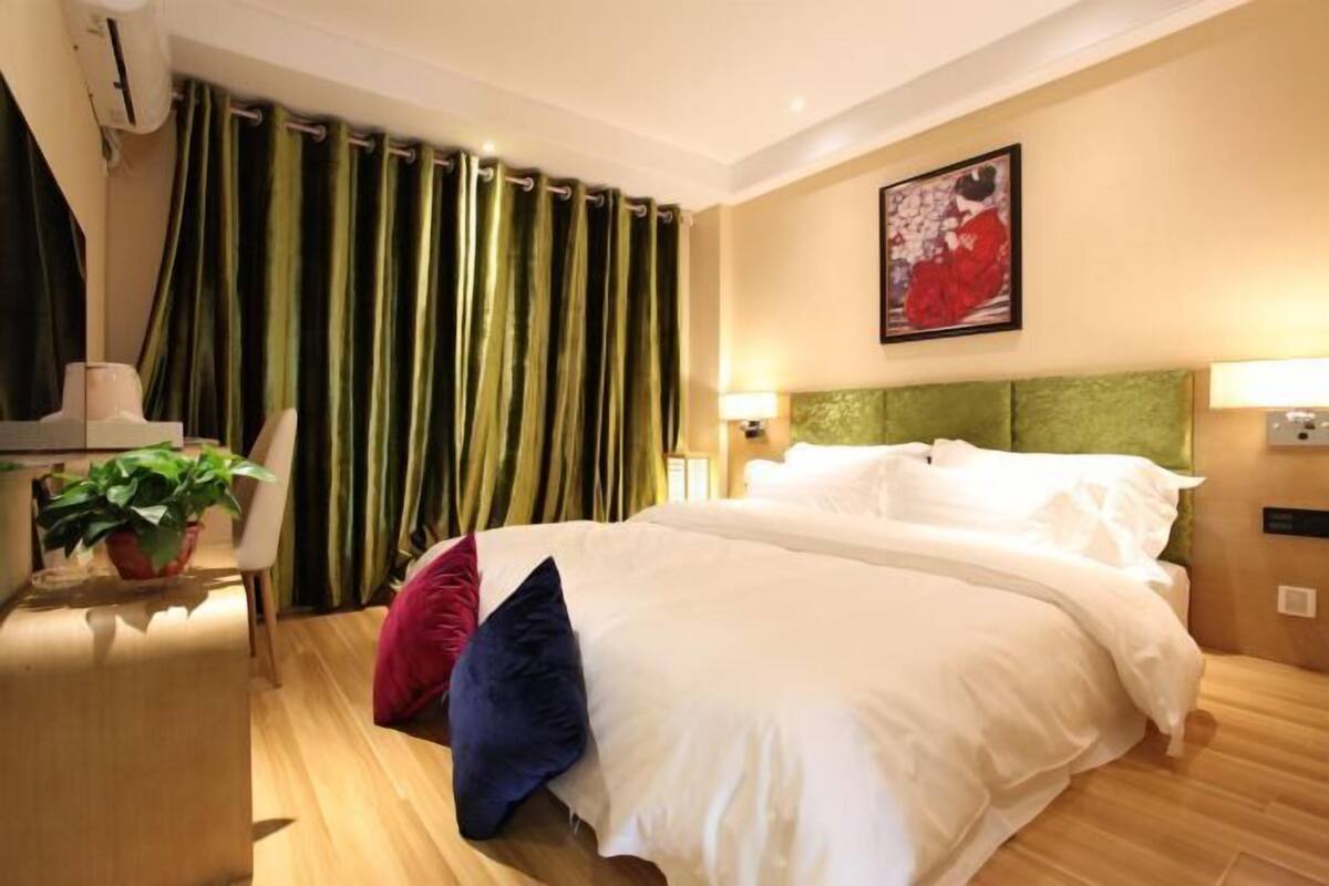 A hotel room with a bed, curtains, a painting, a plant, and a desk.