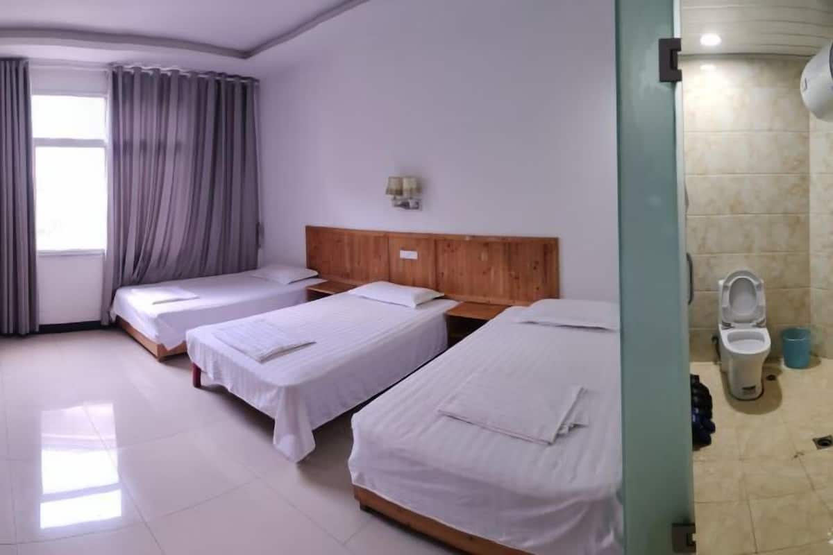 A hotel room with three single beds, a wooden headboard, a window with curtains, a wall-mounted light, and a bathroom with a toilet and sink.
