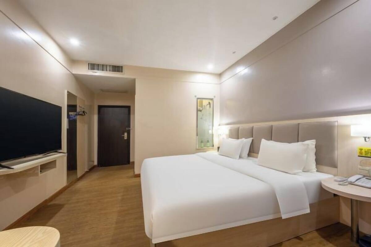 A hotel room with a large bed, a bedside table, a TV mounted on the wall, and a door leading to another room.