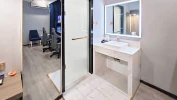 A modern bathroom with a white vanity, a large mirror, and a glass door leading to a seating area with a blue chair and a desk.