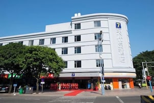 Un hotel blanco con un número '7' destacado en la fachada, una alfombra roja en la entrada y un cielo azul despejado al fondo.