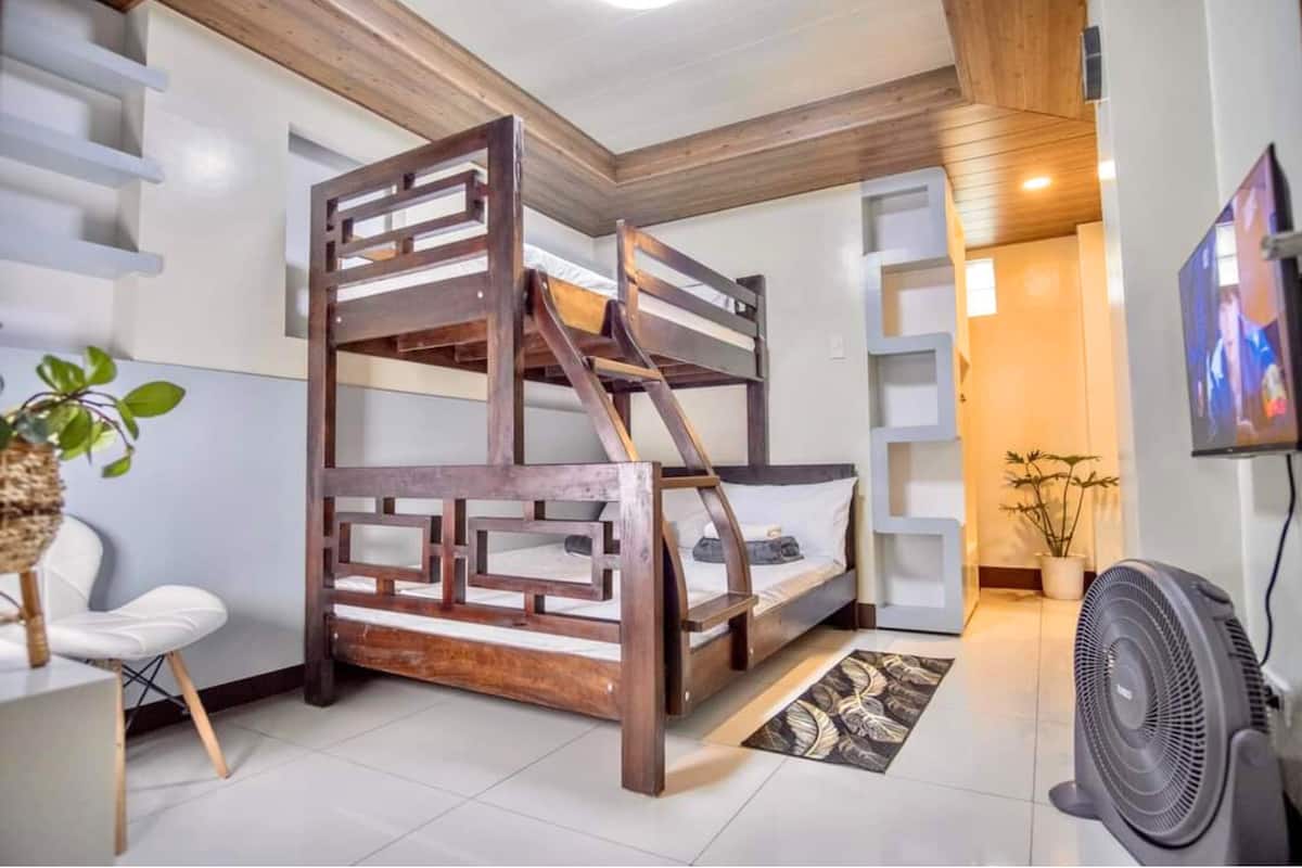 Una cama litera con estructura de madera y ropa de cama blanca, una silla blanca, una planta en maceta, un ventilador y un televisor en la pared.