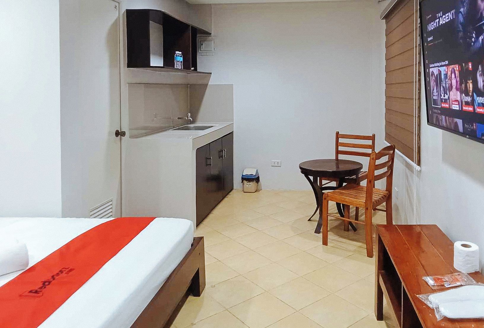Ein kompaktes Hotelzimmer mit Bett, einer kleinen Küchenzeile, einem Esstisch mit Stuhl und einem an der Wand befestigten Fernseher.