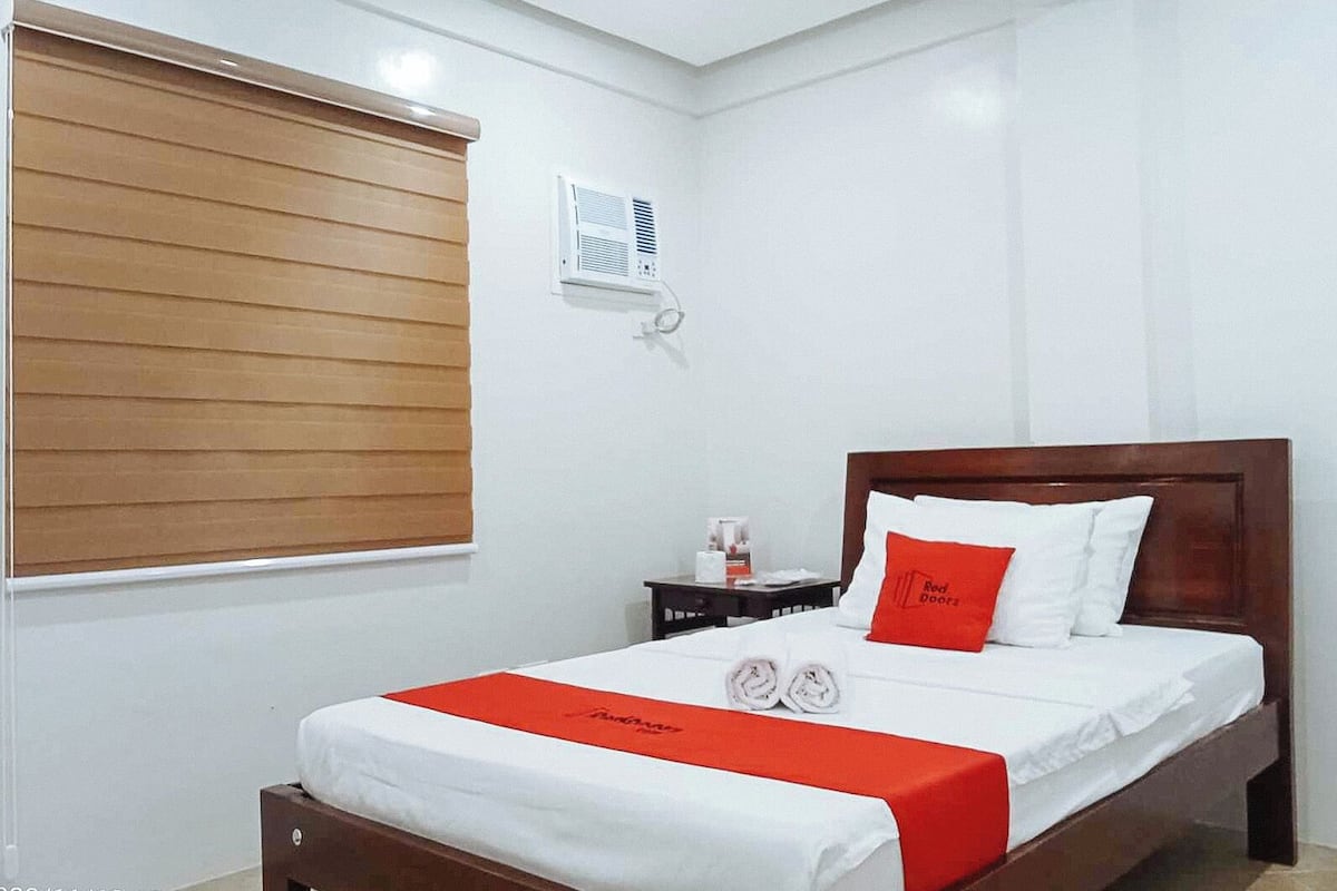 Una cama bien hecha con ropa de cama blanca y roja, un cabecero de madera y una mesita de noche con una taza y pañuelos.