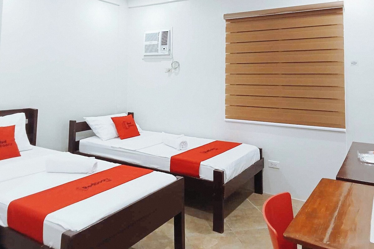 Habitación de hotel con dos camas individuales, un escritorio de madera, una silla, una ventana con persianas y un aire acondicionado instalado en la pared.