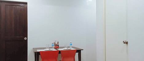 A small room with a wooden door, a white wall, and a table with two red chairs. There are water bottles on the table.