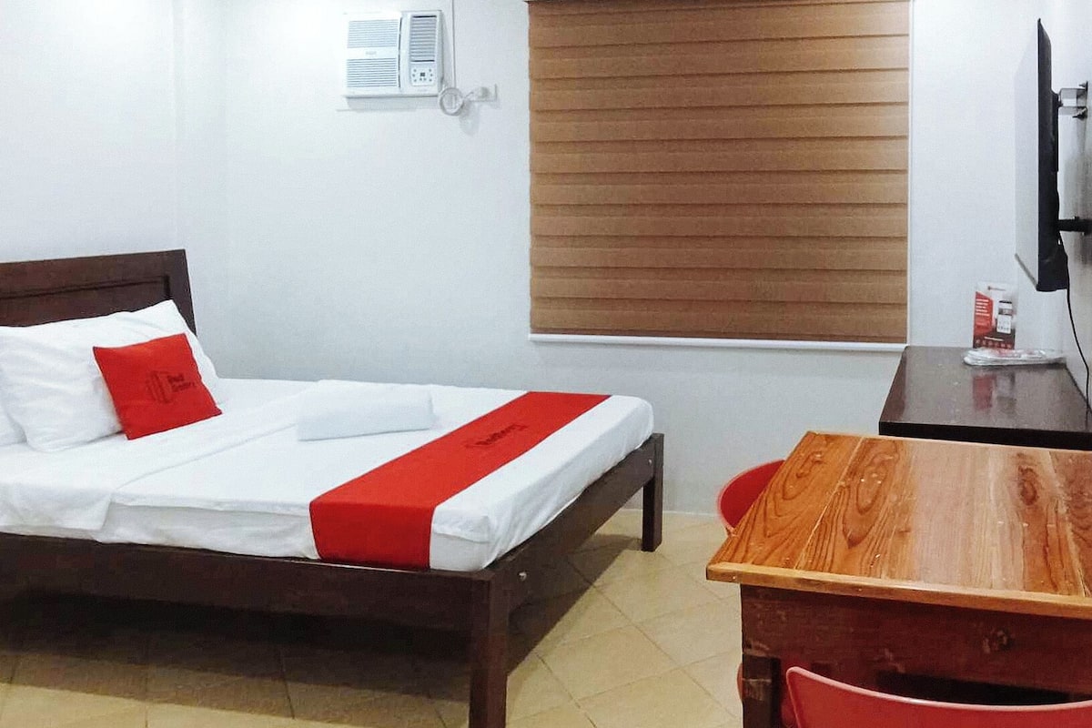 Habitación de hotel con cama, escritorio de madera, una silla, ventana con persianas y aire acondicionado instalado en la pared.