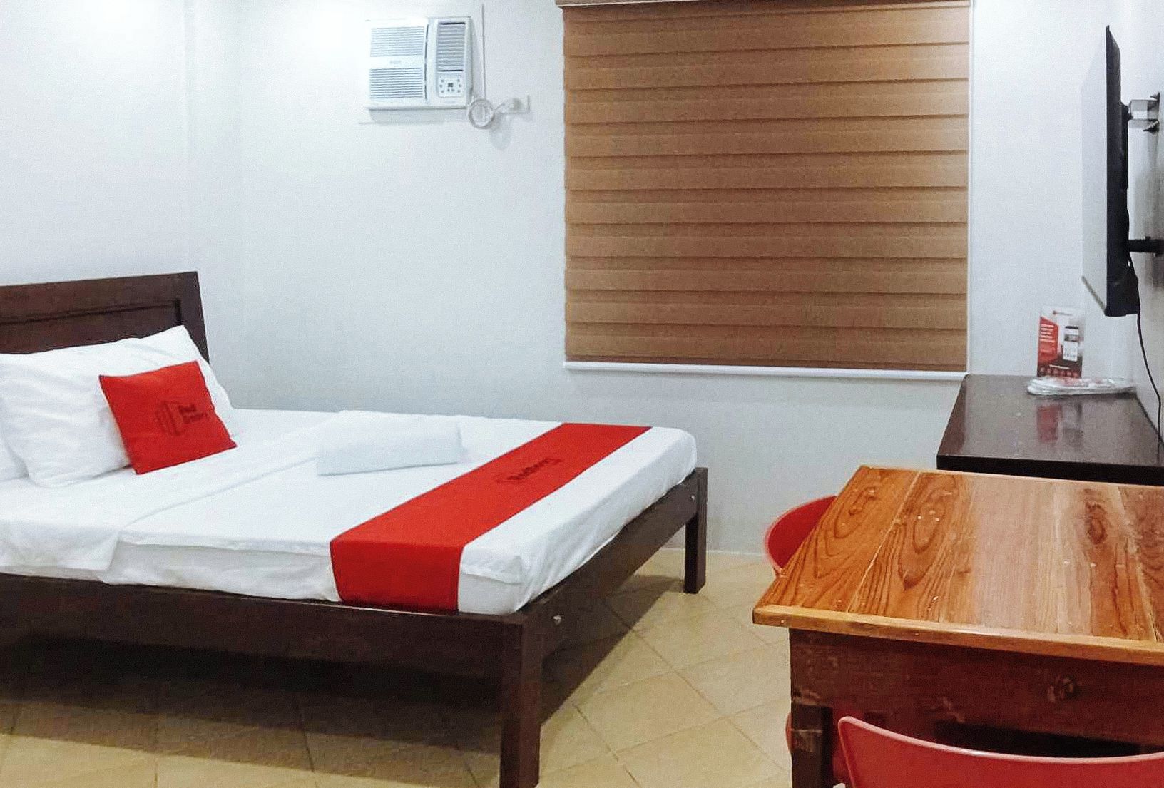 A hotel room with a bed, a wooden desk, a chair, a window with blinds, and a wall-mounted air conditioner.