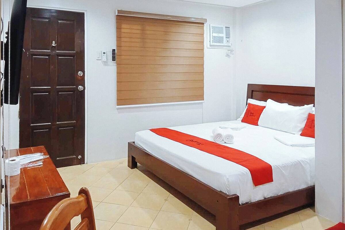 Una habitación de hotel con una cama de madera, ropa de cama blanca, un detalle en rojo, un escritorio de madera y una silla.