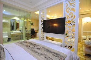 Une chambre d’hôtel dotée d’un grand lit, d’un téléviseur à écran plat fixé sur un mur décoratif, d’une salle de bain avec des parois en verre et d’un coin salon avec un fauteuil.