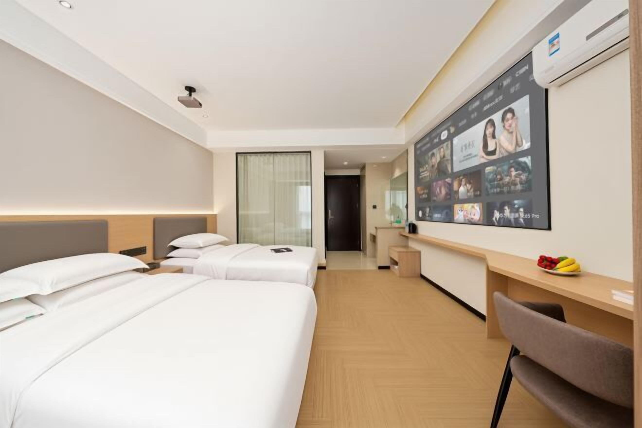 A hotel room with two beds, a desk, a chair, and a TV displaying various channels.