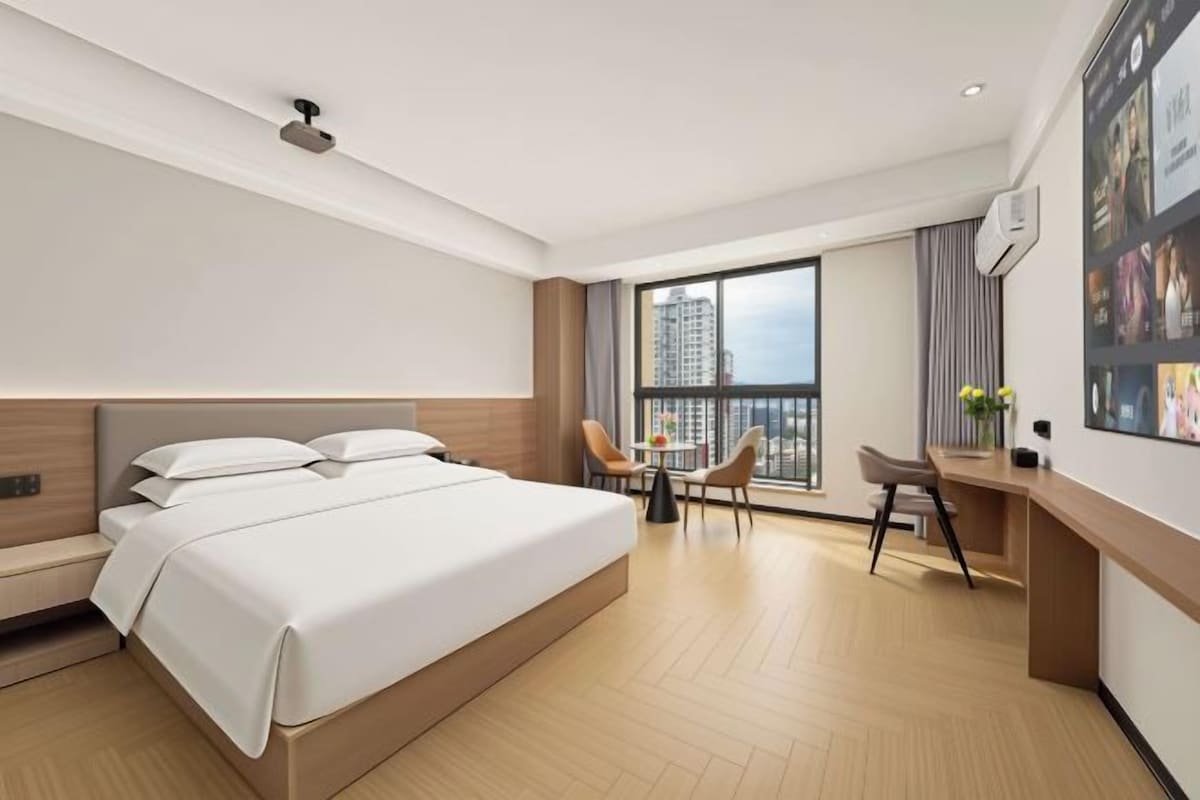 A modern hotel room with a large bed, a desk, and a balcony with a city view.