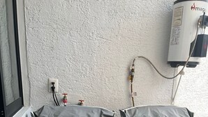 Deux matelas recouverts dans une pièce avec un mur blanc à la texture particulière, une fenêtre et un chauffe-eau.