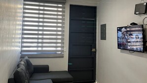 Un petit salon avec un canapé de couleur sombre, un téléviseur fixé au mur et une fenêtre équipée de stores.