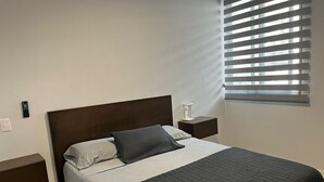 Un lit bien fait, avec une tête de lit sombre, une couvre-lit grise et noire et un oreiller. On y trouve une table de chevet sur laquelle est posée une lampe.