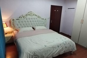 Een slaapkamer met een gestoffeerd hoofdeinde, een bed met een bloemenprint dekbedovertrek, een nachtkastje met een lamp en een kledingkast.