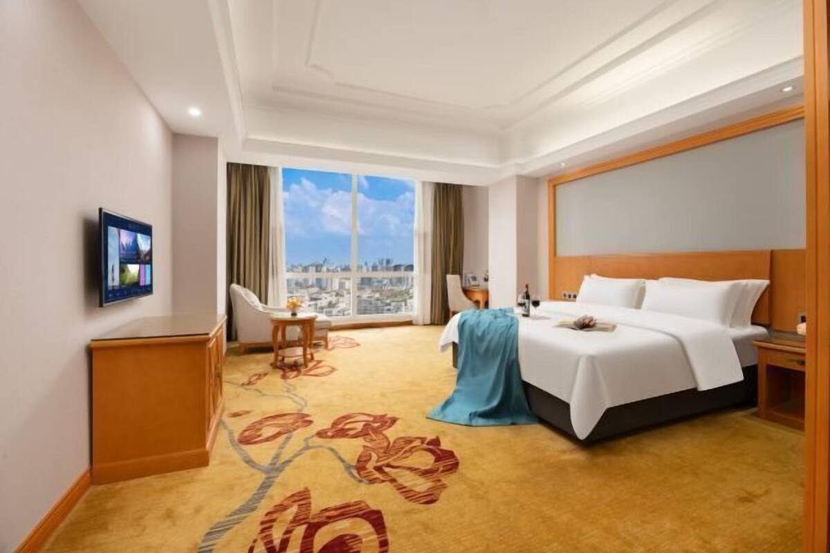 Ein Hotelzimmer mit Bett, Schreibtisch, Stuhl, Fernseher und einem großen Fenster mit Blick auf die Stadt.