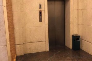 An elevator with a control panel and a trash bin in a hallway with tiled walls. - Jinyuwan Hotel (Dalian)