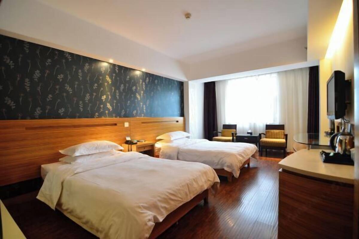 A hotel room with two beds, a wooden headboard, a desk with a chair, a TV, and a large window with curtains.