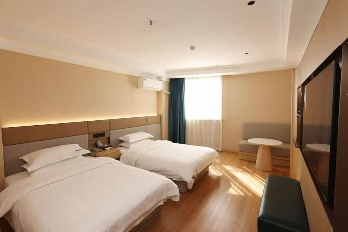 A hotel room with two beds, a small round table, a bench, and a large window with curtains.