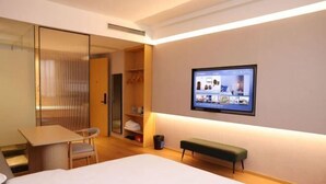 A hotel room with a bed, a desk, a chair, a bench, a TV mounted on the wall, and a closet with hanging clothes. - Ruihao Hotel (Pingyang)
