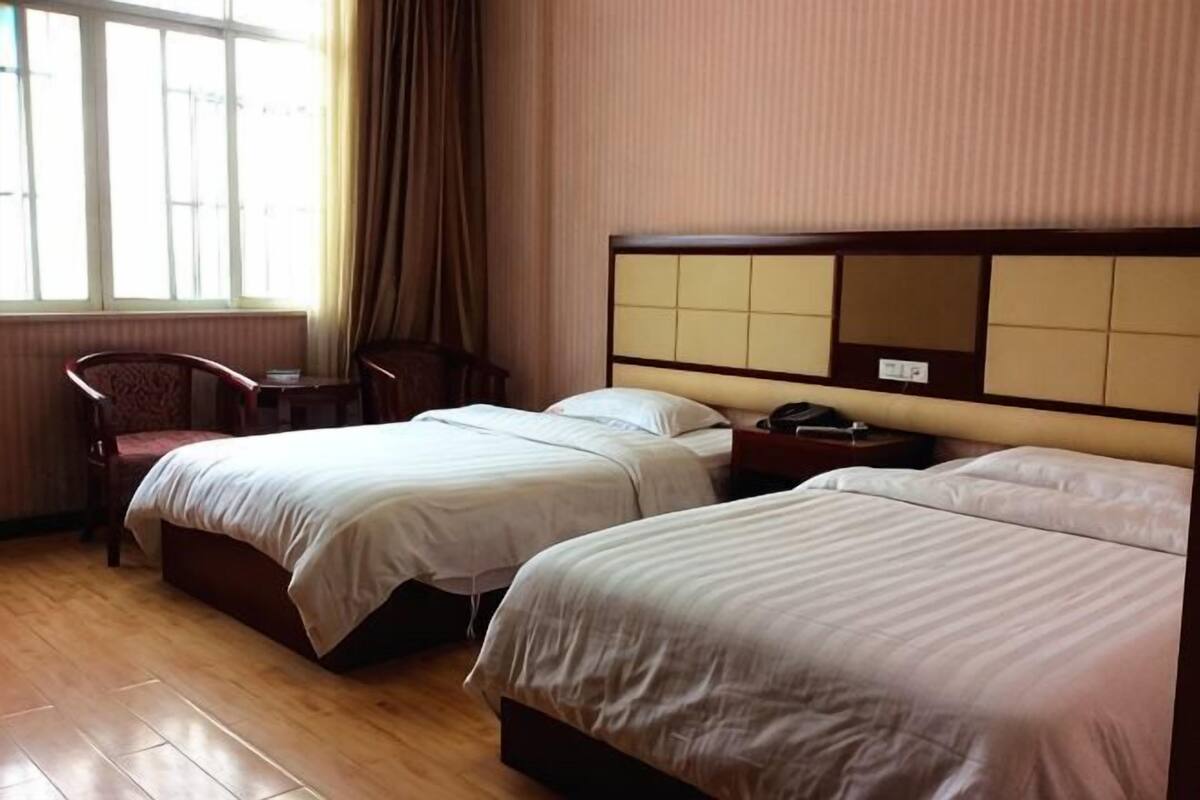 A hotel room with two beds, a chair, a bedside table, and a window with curtains.