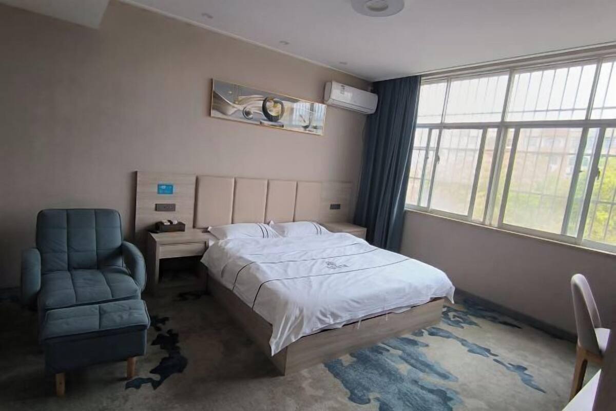 A hotel room with a bed, a chair, a nightstand, a painting, and a window with curtains.