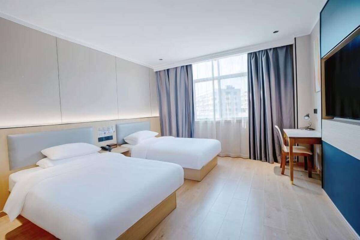 A hotel room with two beds, a desk, a chair, a TV, and a window with curtains.