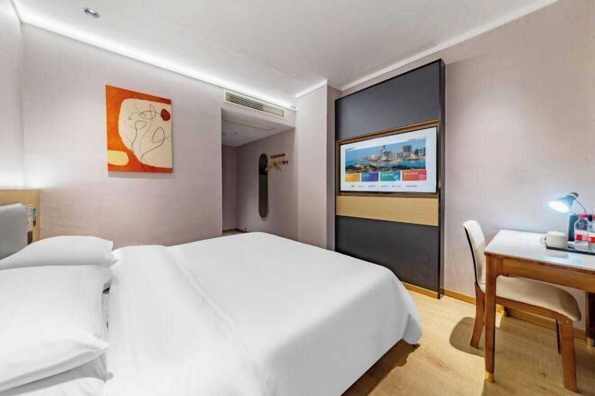 A hotel room with a bed, a desk with a chair, a TV, and a painting on the wall.