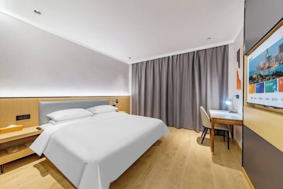 A modern hotel room with a bed, desk, and chair, a large window with curtains, and a wall-mounted TV.