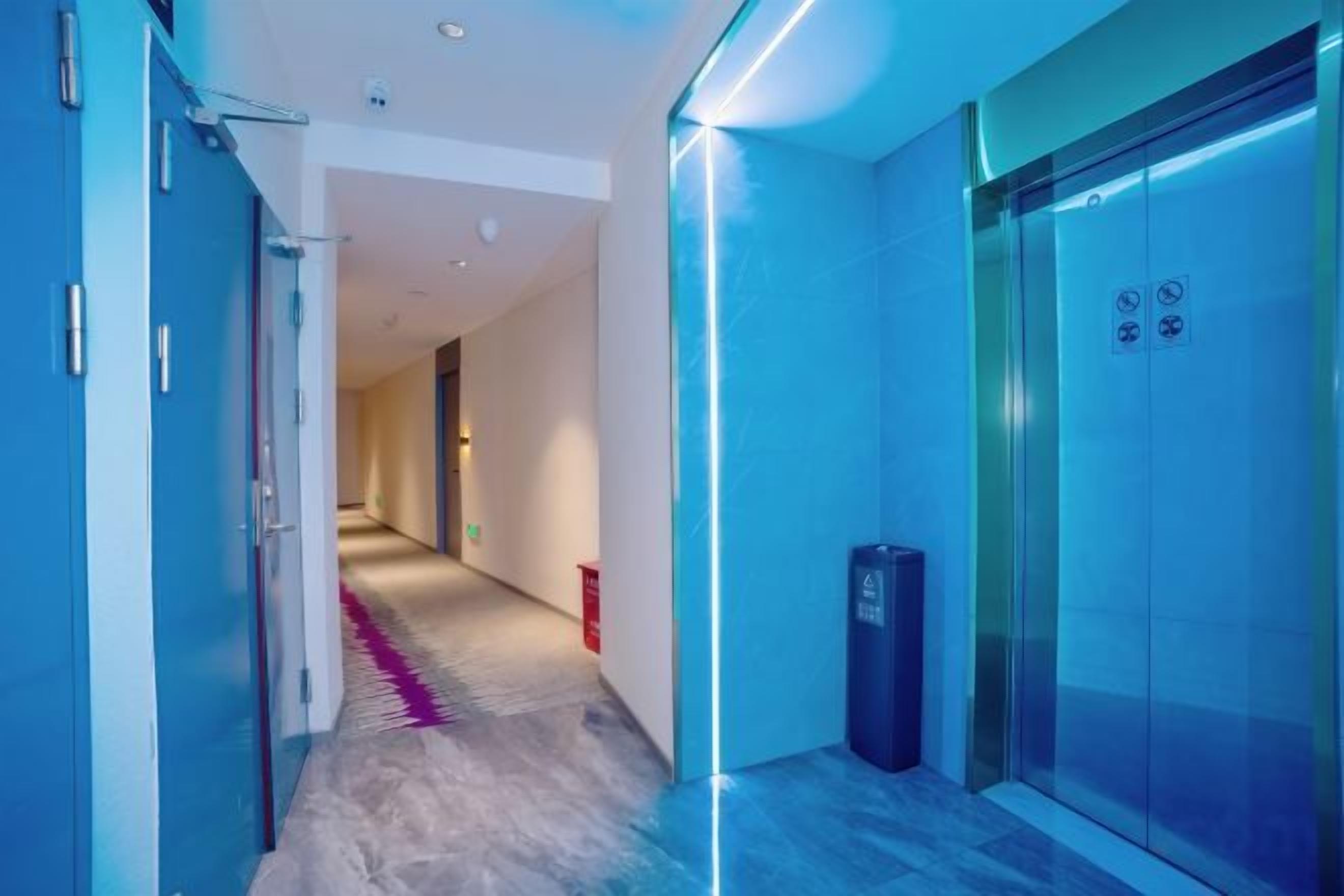 A corridor with a glass elevator and a trash bin, leading to a room with a door.