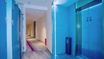 A corridor with a glass elevator and a trash bin, leading to a room with a door.
