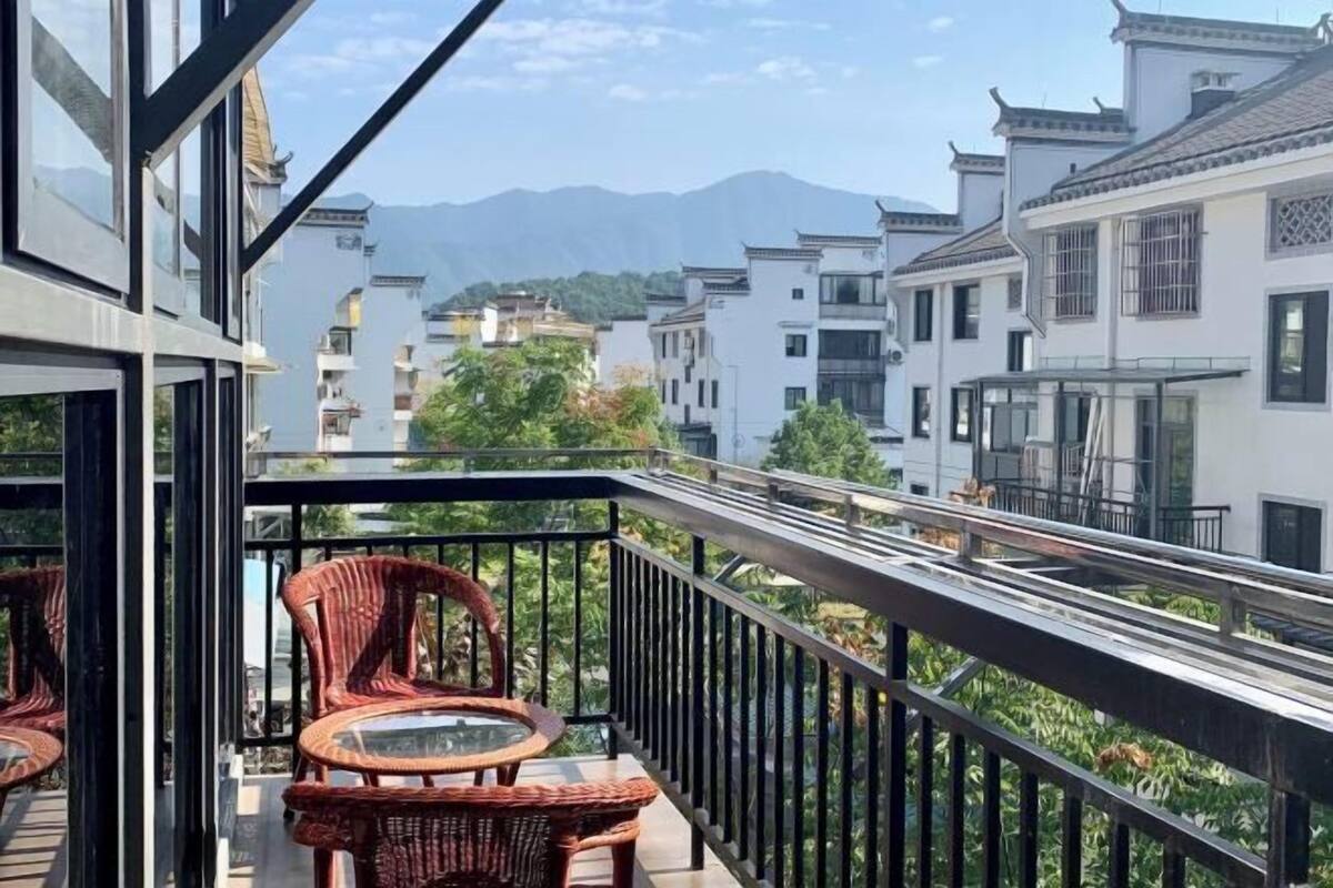 Un balcón con mobiliario de mimbre y una mesa de vidrio, con vistas a edificios tradicionales y una cadena montañosa.