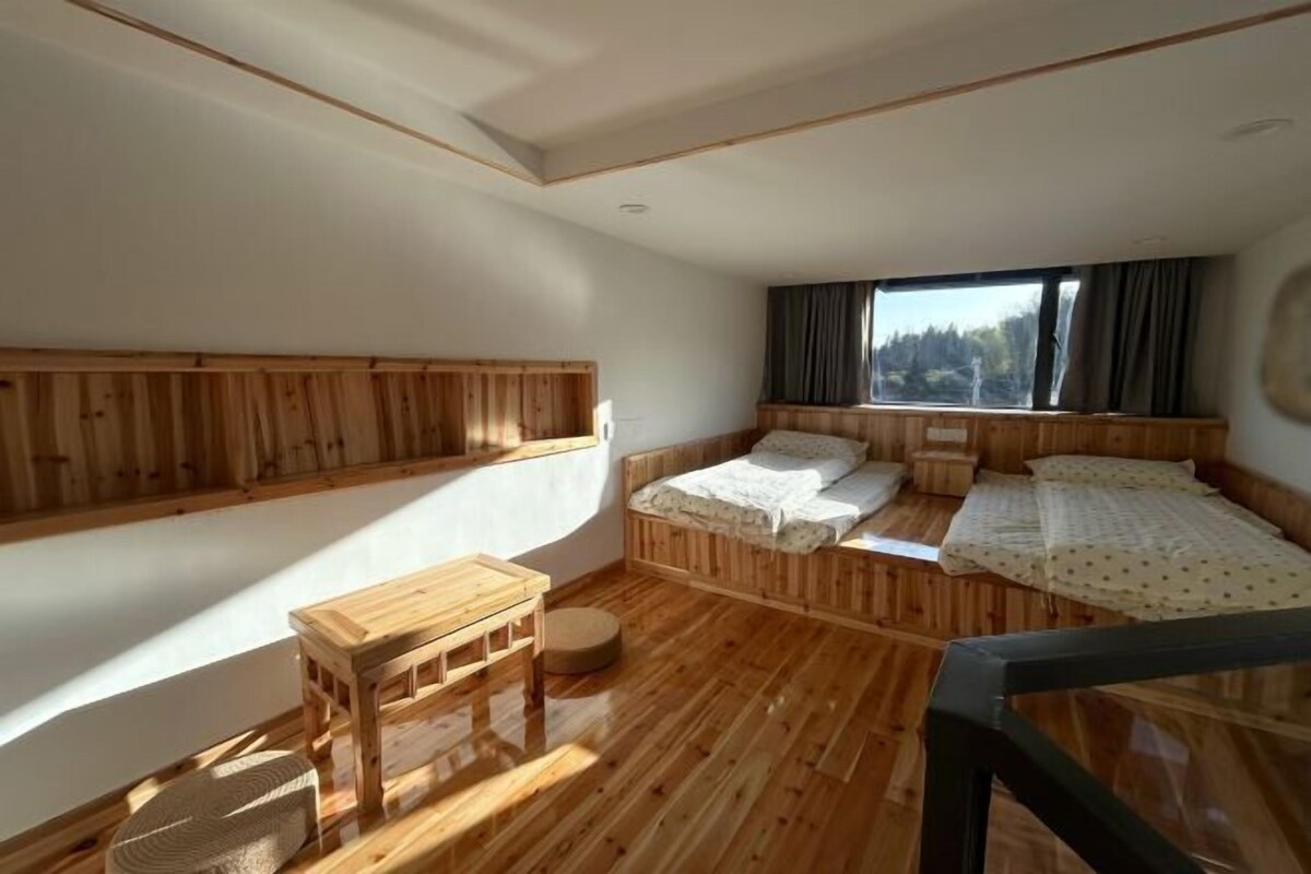 Una habitación con suelo de madera, dos camas, una mesita y un ventanal con cortinas.